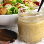 Caesar dressing in a glass jar with a white bowl of salad