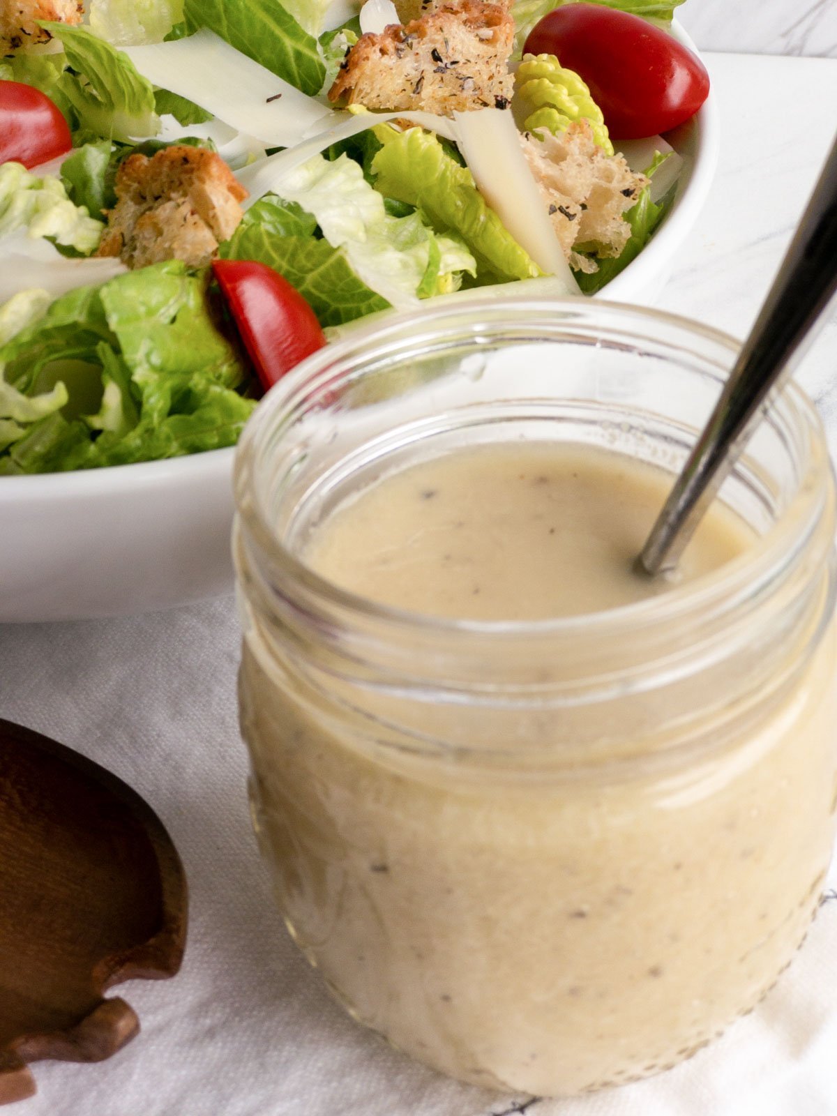 Caesar dressing in a jar with a spoon in it and a salad next to it.