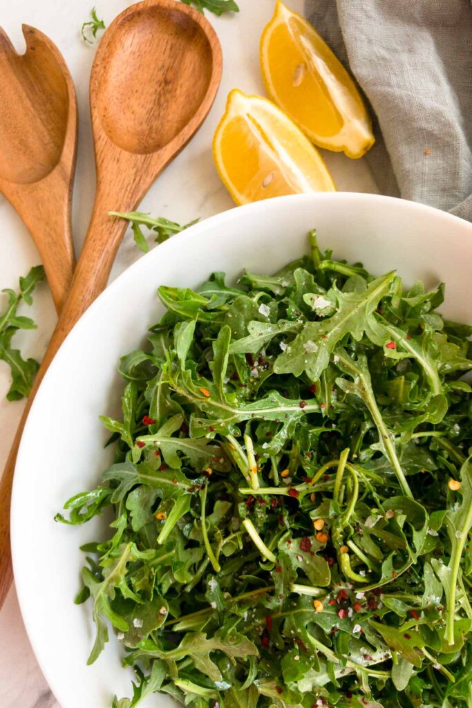 Arugula in a white bowl with lemons and salad tongs on the side