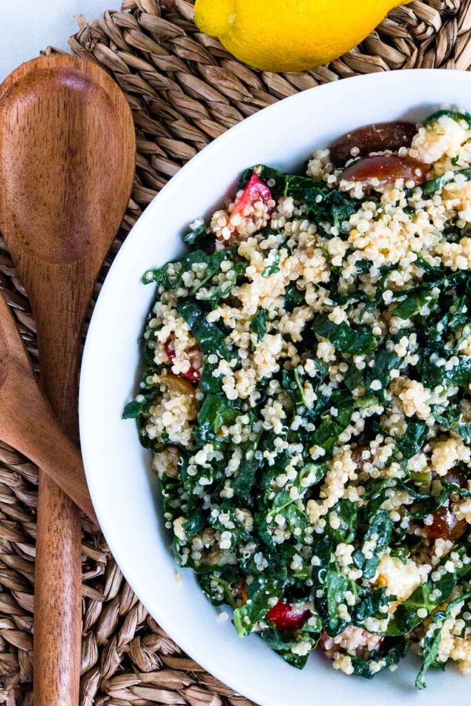 Kale and quinoa salad with tomatoes in a bowl, wood salad tongs next to bowl and lemon above bowl