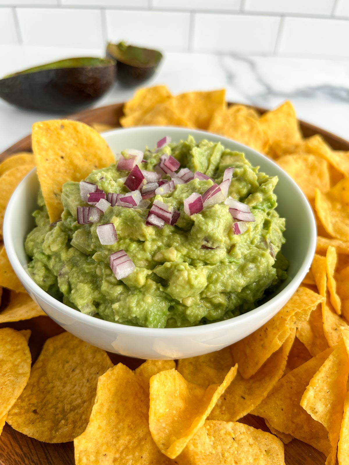 Guacamole with diced red onion garnish, single chip in guacamole in a white bowl surrounded by tortilla chips 