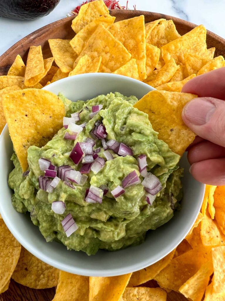 Hand with a chip dipping into a white bowl filled with guacamole.  