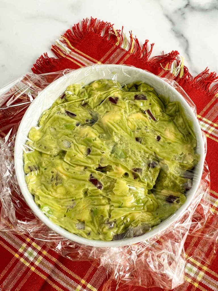 Guacamole in a white bowl covered with plastic wrap on a rust colored napkin