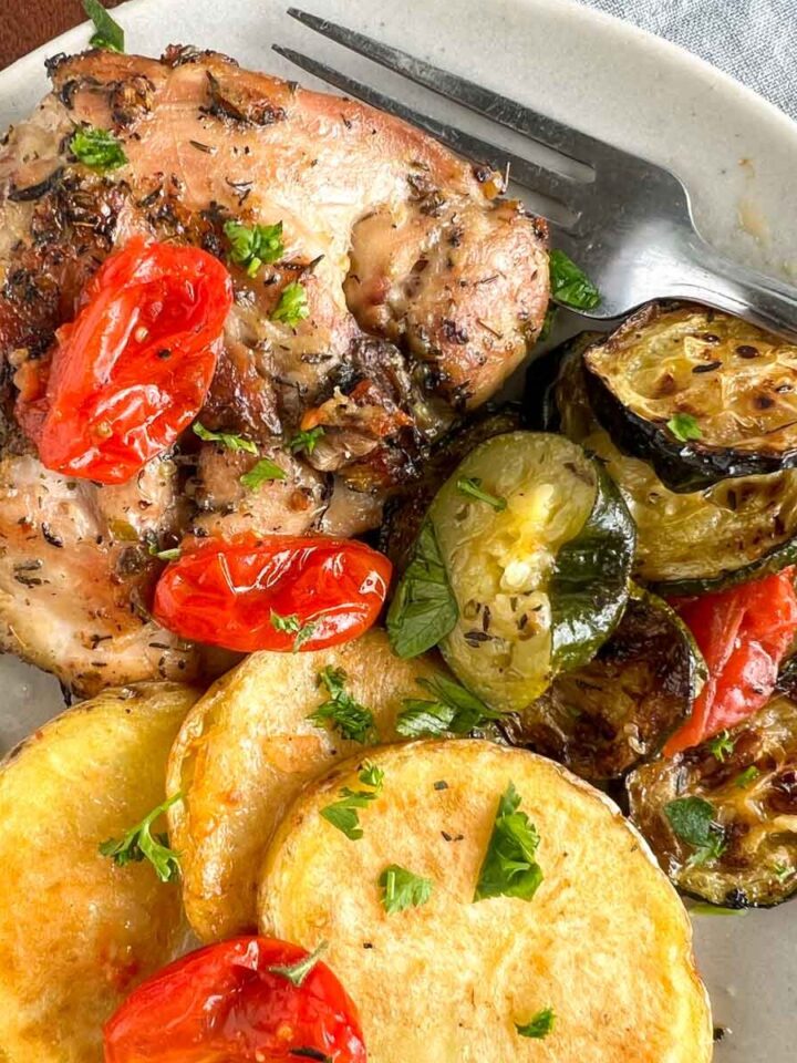 Baked Chicken thigh with roasted zucchini and potato rounds with cherry tomatoes on a white plate