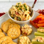 Pimento cheese in a white bowl, surrounded by crackers, pimento topped crackers, pimento stuffed celery and baby carrots