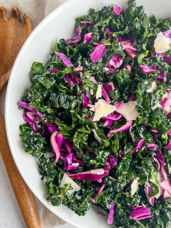 Shredded Kale and chopped red cabbage in a white bowl with wood tongs on the left side