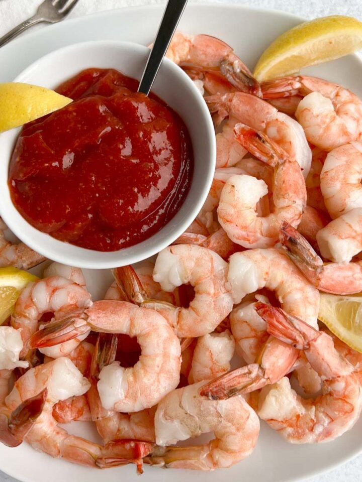 Cooked shrimp with lemon wedges on a white square plate with cocktail sauce in a round bowl next to the shimp