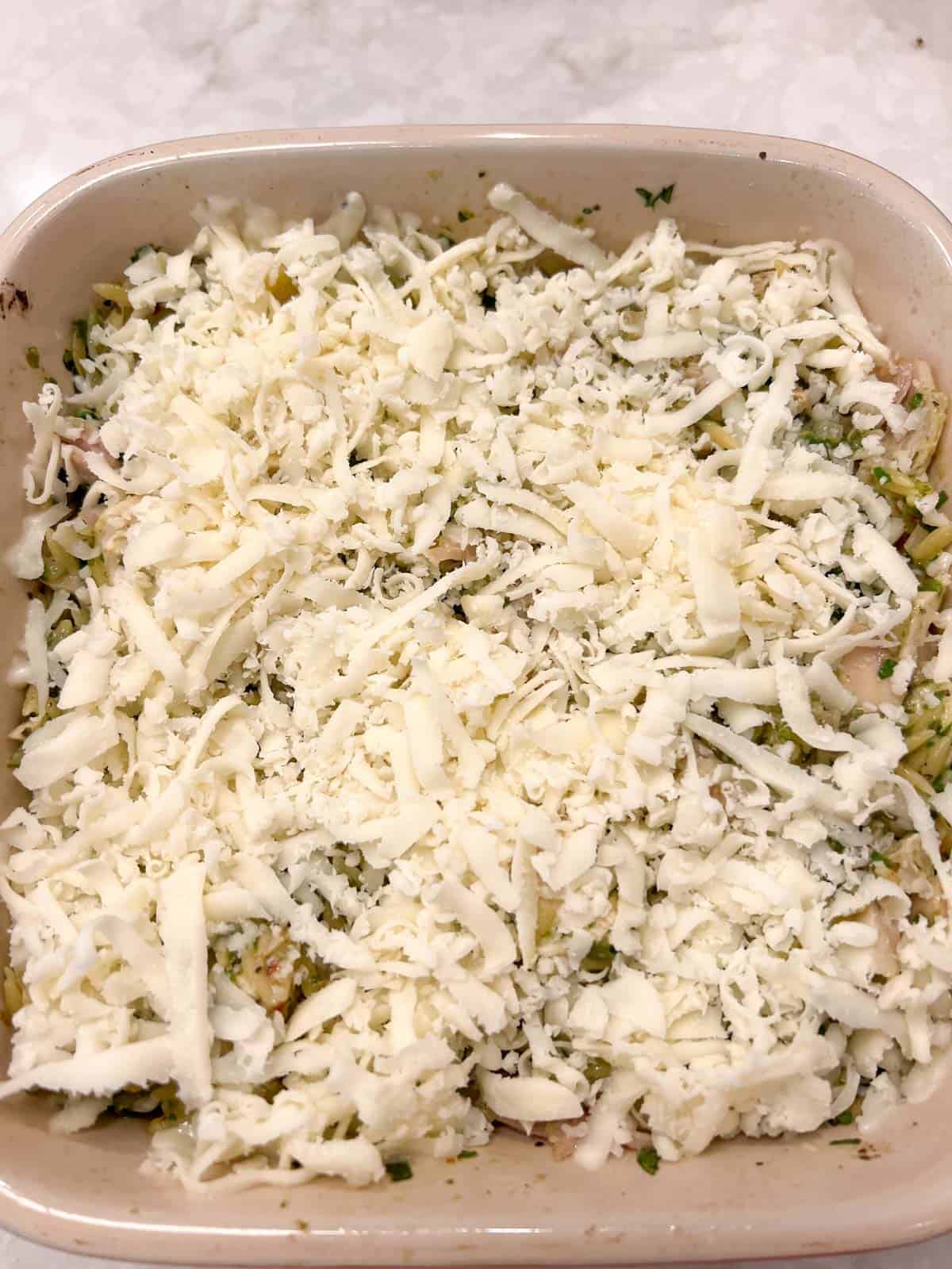 Shredded mozzarella cheese on chicken, orzo, spinach and pesto in a casserole dish