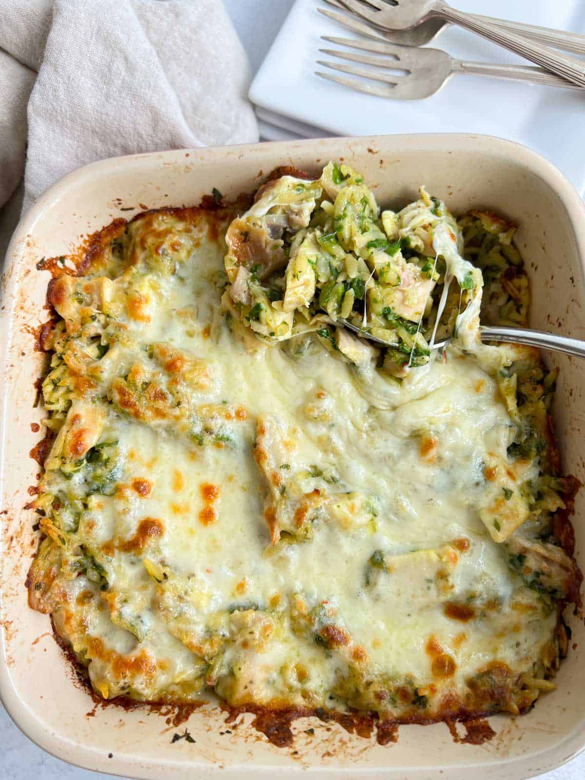 Serving spoon with chicken, orzo and pesto in a square baking pan.