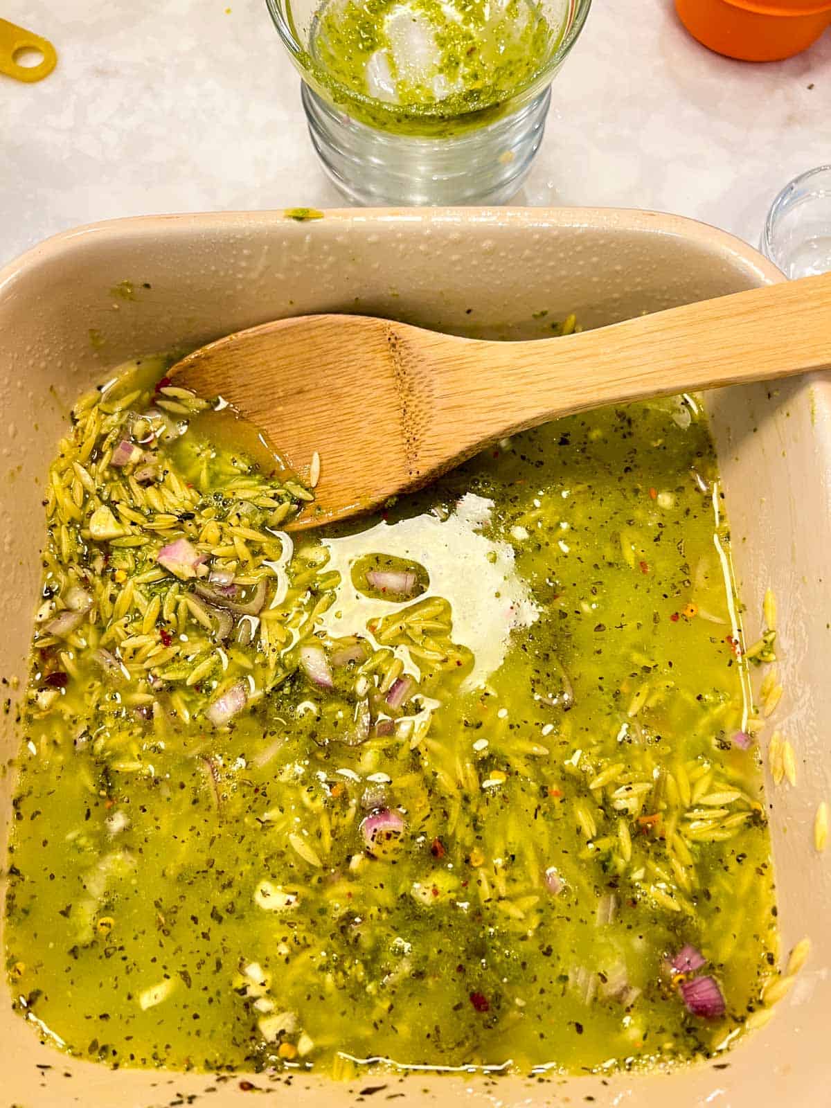  liquid ,orzo and pesto in a casserole dish with a wooden spoon