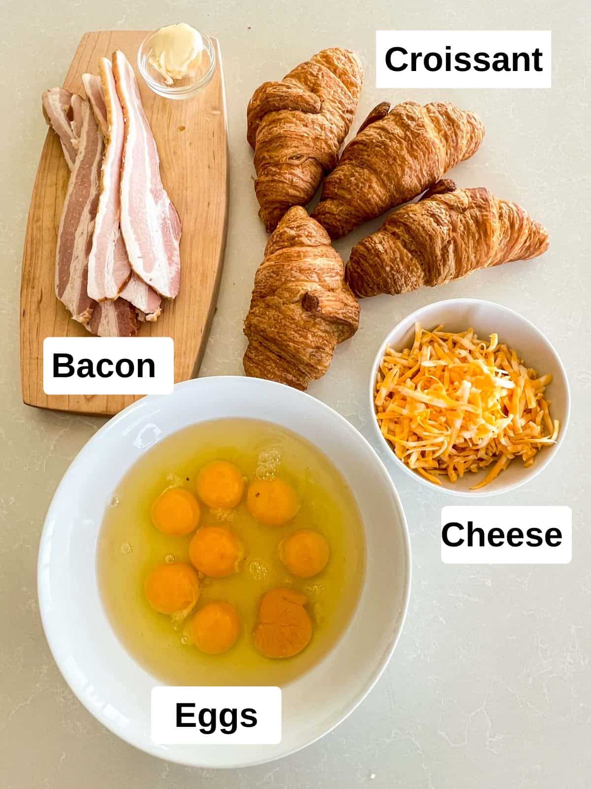 Bacon on a wood board, croissants, eggs cracked into a white bowl, and grated Colby Jack cheese in a white bowl.
